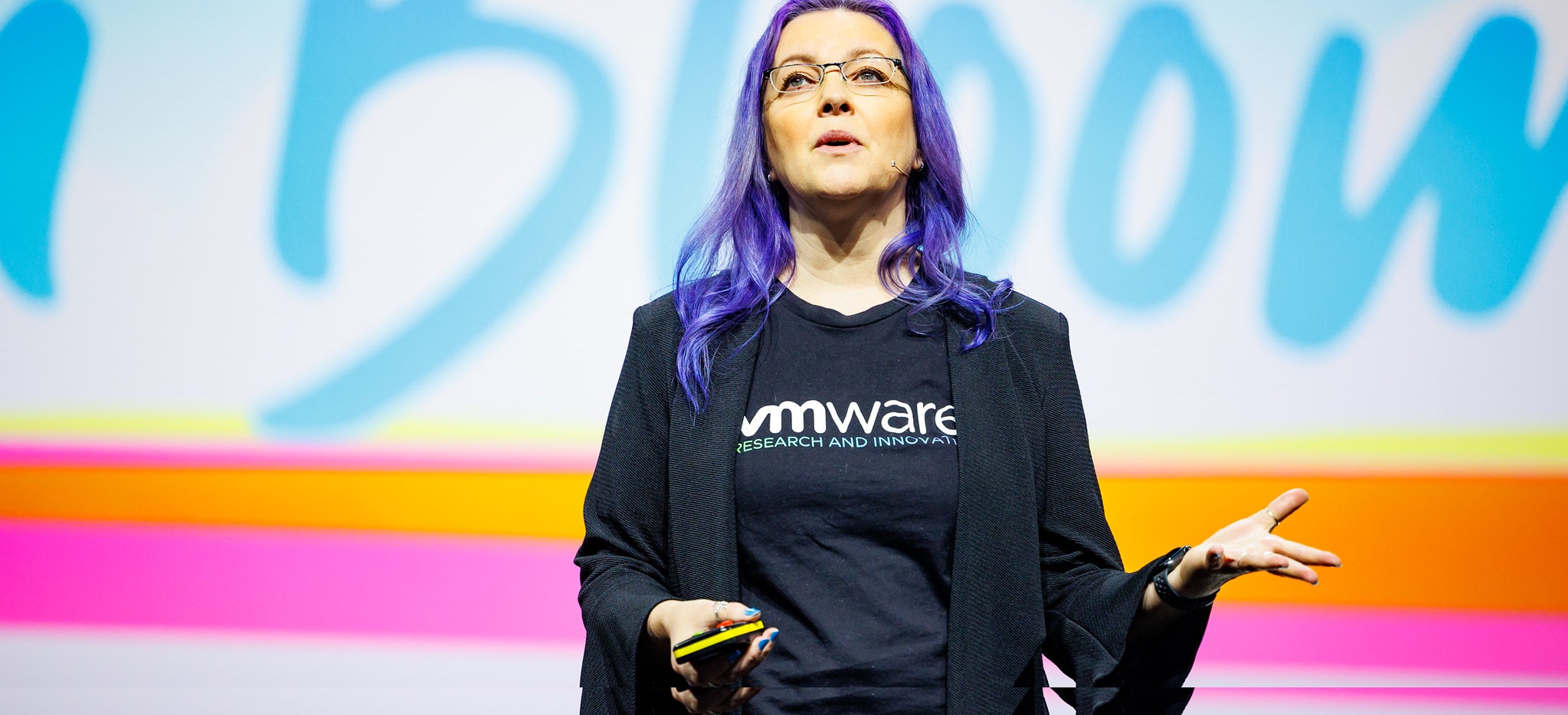 Dawn giving a keynote at KubeCon EU wearing all black with a VMware t-shirt and purple hair in front of colorful background.