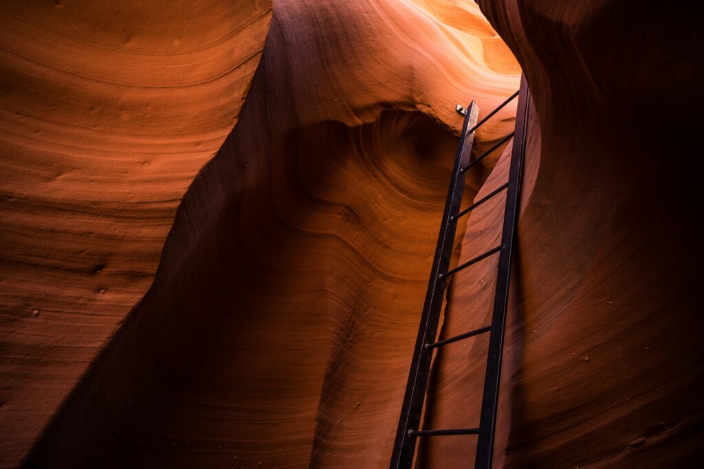 Black ladder going through a slot in a canyon with reddish orange rock.
