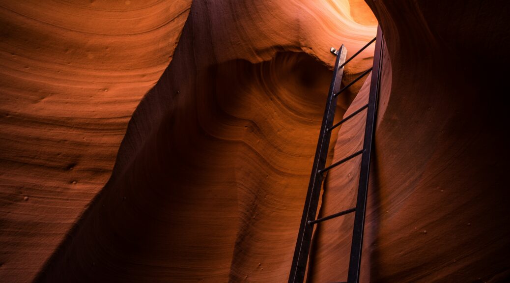 Black ladder going through a slot in a canyon with reddish orange rock.