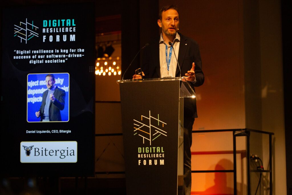 Daniel Izquierdo on the stage behind a podium with a Digital Resilience Forum sign. On the left is a screen with Daniel's picture and the Bitergia logo reading "Digital resilience is key for the success of our software-driven digital societies"