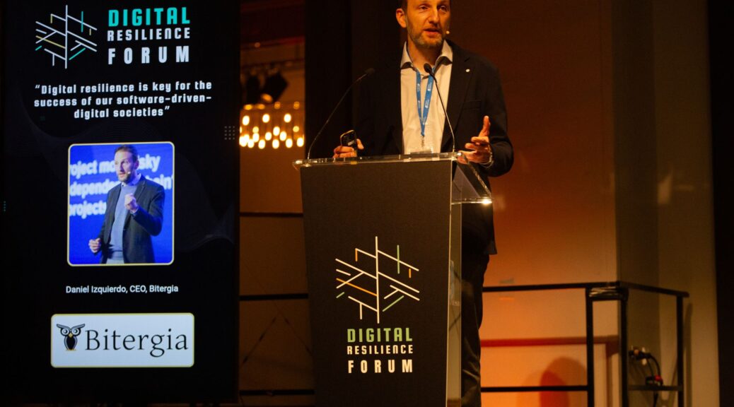 Daniel Izquierdo on the stage behind a podium with a Digital Resilience Forum sign. On the left is a screen with Daniel's picture and the Bitergia logo reading "Digital resilience is key for the success of our software-driven digital societies"