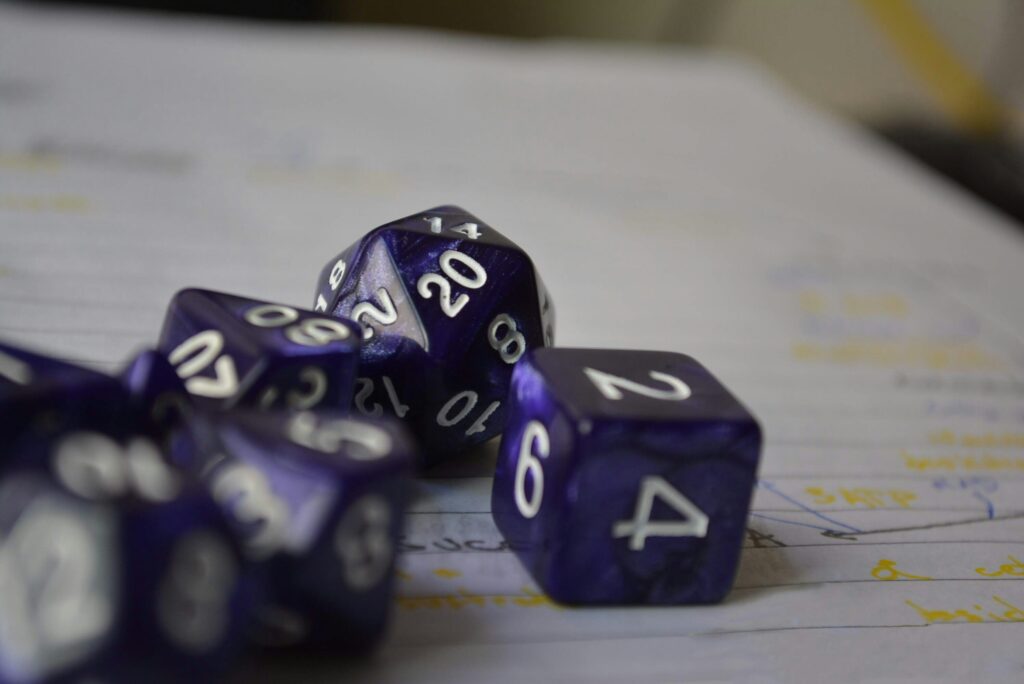 Various types of purple dice with white letters, including a traditional D6 die, a D20, and others. Sitting on a white piece of paper with blurred writing.