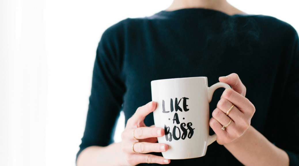 Woman in a black shirt standing in front of a white background holding a white mug reading, "Like A Boss"
