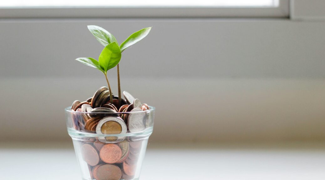 Tiny green plant growing out of a glass cup filled with various coins.