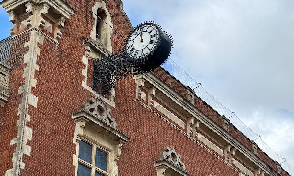 Black clock with a white face at 12 attached via ornate black metal to the side of a red brick building that is an old town hall in an English village.